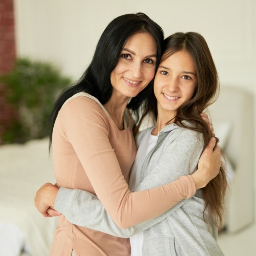 Adult and young person smile at the camera while hugging closely in a bright room with neutral furnishings.