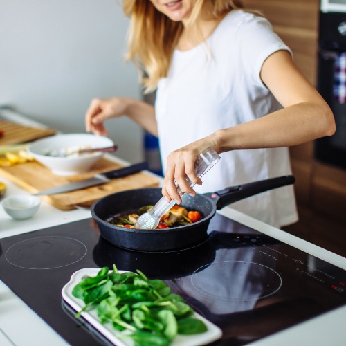 Woman cooks vegetables in a frying pan at a kitchen island with cucumbers, tomatoes, greens, and bowls nearby.