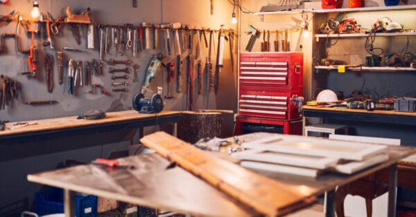 Busy workshop features rows of hanging tools, a red storage chest, wooden worktables, and equipment on open shelves.