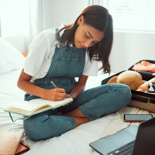 A young woman sits on the end of her bed with a suitcase next to her as she writes in a notebook all her travel plans.