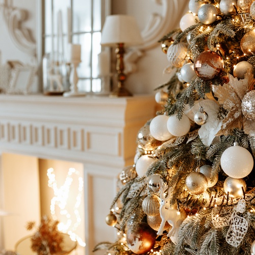 Shimmering metallic ornaments, white baubles, and white lights adorn a Christmas tree that's next to a white fireplace.
